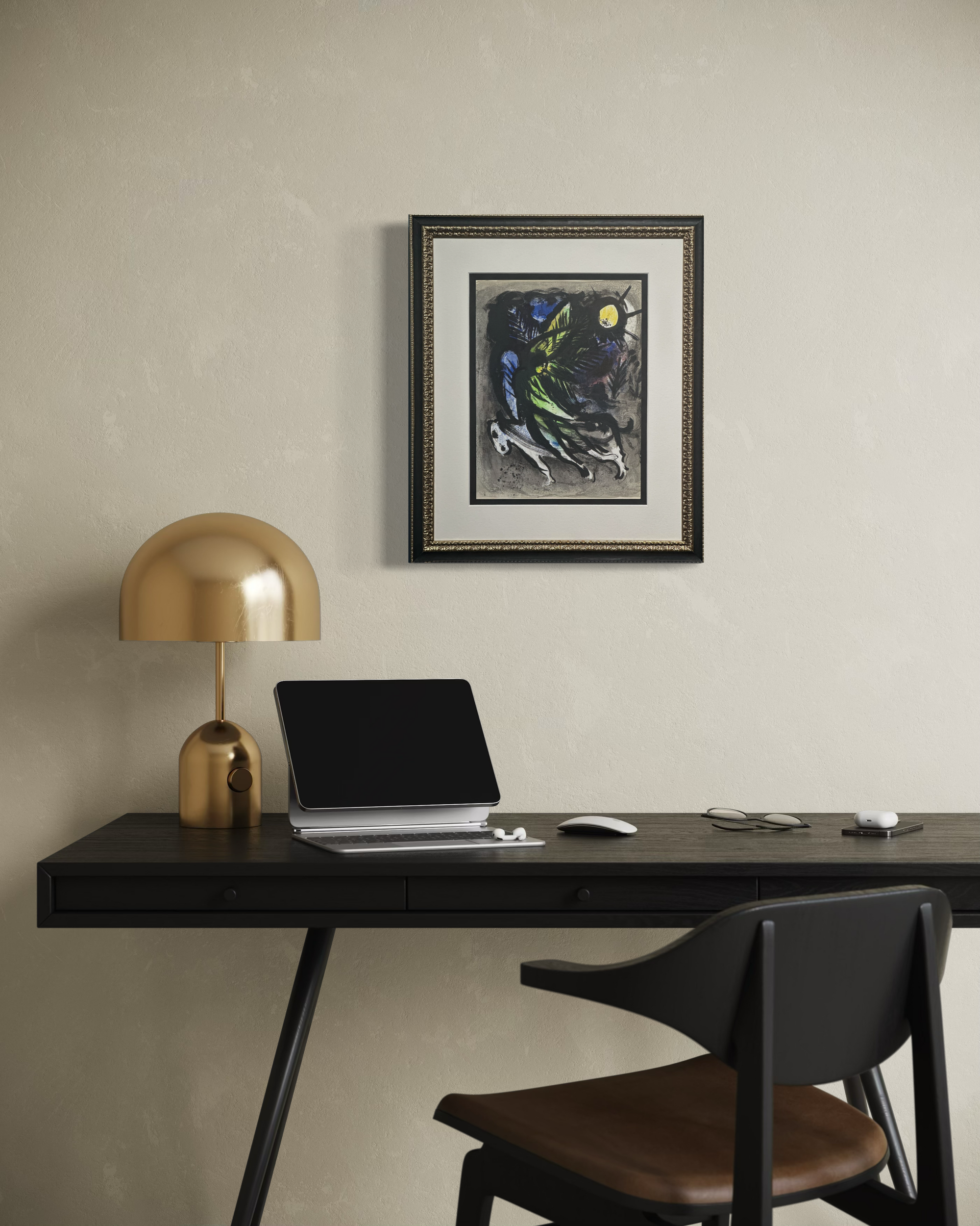 A framed abstract art print hangs on a textured beige wall above a dark wood desk setup with a gold dome lamp, laptop, mouse, and chair.