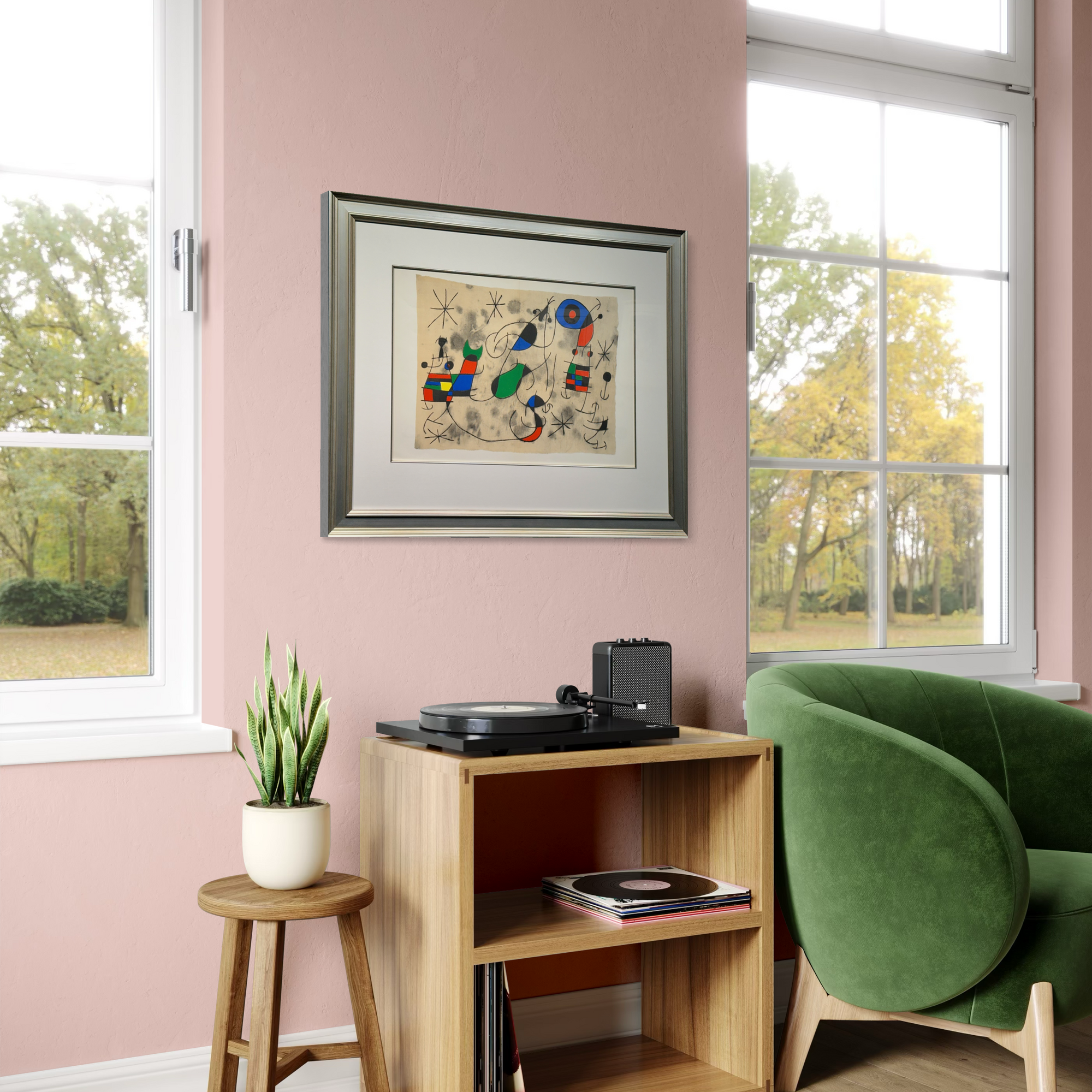 A framed abstract art print with a colorful composition and a gray frame hangs on a dusty pink wall in a styled room. The room features a light wood side table with a turntable, a small potted plant on a wooden stool, and a green velvet armchair next to a window.