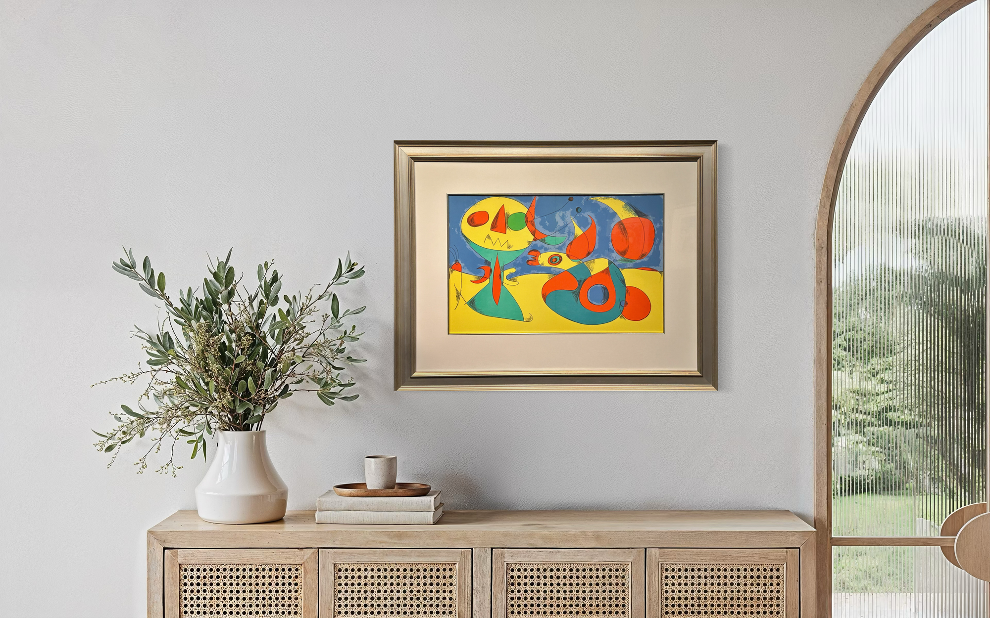 A framed abstract art print with bold yellow, red, and blue shapes hangs on a light gray wall above a wooden cabinet, styled with a white vase of greenery and a small cup.