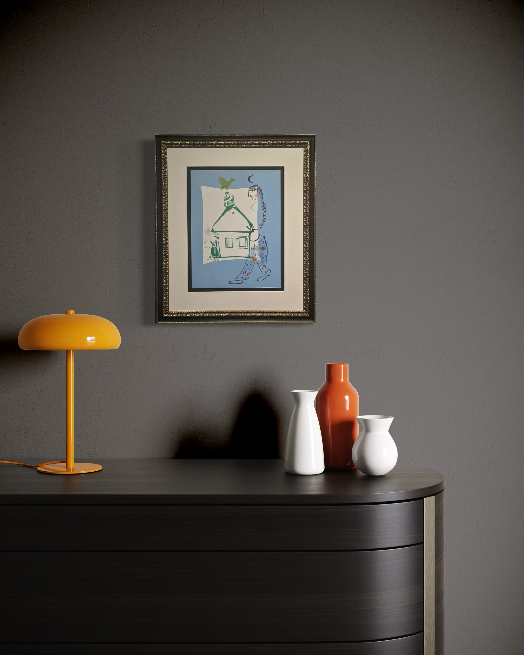 A framed art print featuring a whimsical blue house illustration hangs on a dark gray wall above a dark wood console table. The table is styled with a bright yellow table lamp on the left and three ceramic vases in white, orange, and white on the right.
