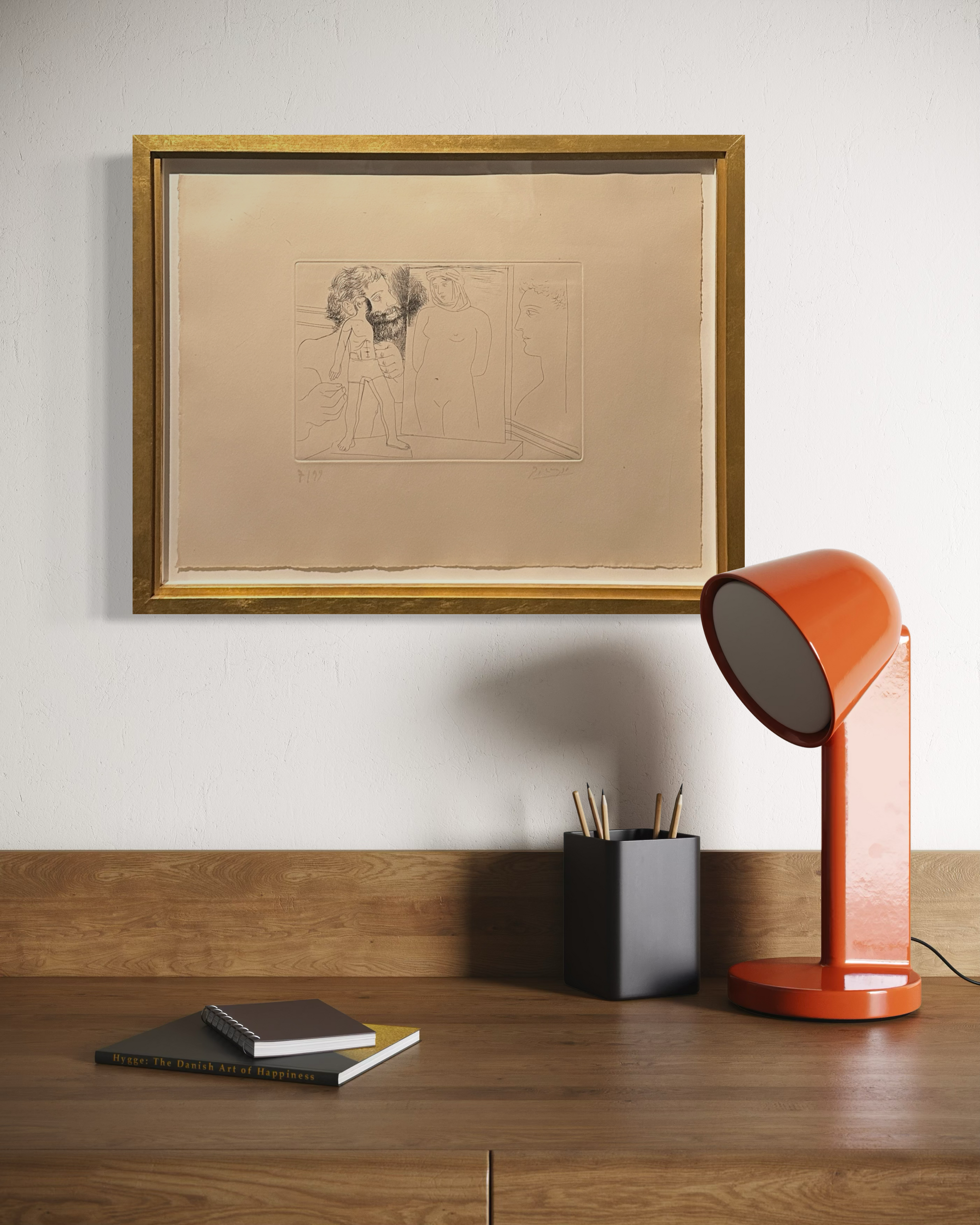 A framed art print with a gold frame hangs on a white wall above a wooden desk. The print features a pencil sketch on beige paper.